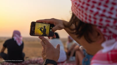 person with red and white head wrap holding cell phone with falconry image, sandy background with other people sitting