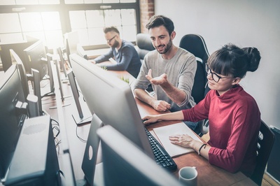 Three people sitting in front of desktop computers talking.