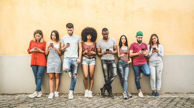 Group of young adults standing against a wall using their cell phones.